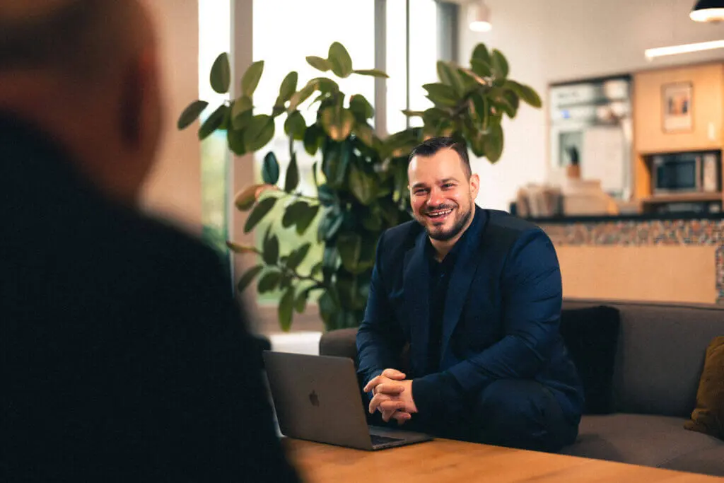 Ein fröhlicher Mann in einem dunkelblauen Anzug sitzt an einem Holztisch und lächelt, während er seinem Gesprächspartner gegenübersitzt und von Maßgeschneiderten Lösungen für PC- und Mac-Rechner erzählt. Im Hintergrund sind große Grünpflanzen und ein helles, modern eingerichtetes Büro mit sichtbarer Küchenzeile.