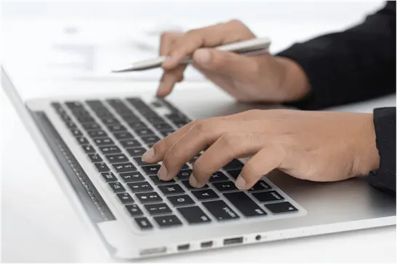 Eine Person tippt auf der Tastatur eines MacBook, während sie in der anderen Hand einen Stift hält, um Notizen zu machen